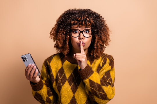 Young woman with curly hair and glasses holding smartphone and making a shushing gesture in a yellow argyle sweater, expressing surprise, secrecy and quiet communication