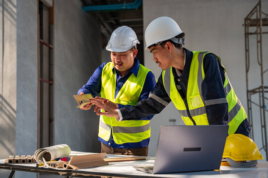 Two Asian construction engineers in safety gear examining and discussing a building material sample at an indoor construction site.