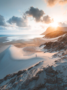 Aerial view of Detwah Lagoon Ramsar Site with a person walking on white sand dunes at sunset, rocky cliffs, and calm sea in Socotra, Yemen.