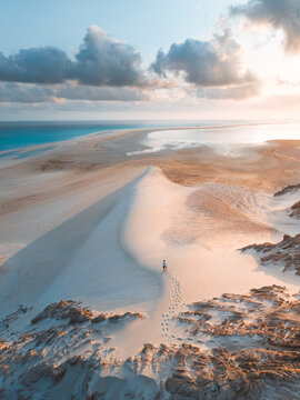 Aerial view of Detwah Lagoon Ramsar Site with a lone person walking across white sand dunes leaving footprints, surrounded by turquoise waters and soft golden light, Socotra, Yemen.