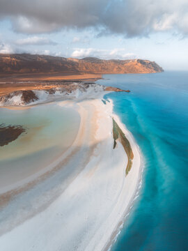 Aerial view of Detwah Lagoon Ramsar Site with white sand beaches, turquoise waters, and mountains under a cloudy sky in Socotra, Yemen.