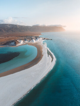 Aerial view of Detwah Lagoon Ramsar Site with its pristine white sandbar separating turquoise waters from the deep blue sea, set against an arid mountainous coastline, Socotra, Yemen.
