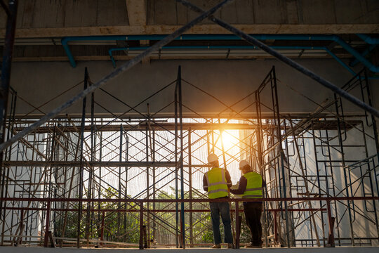 Silhouette of two Asian construction engineers discussing a blueprint inside an unfinished building with scaffolding and bright sunlight.