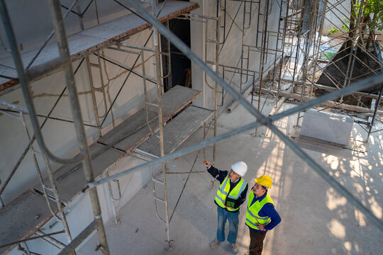 High angle view of two Asian construction engineers in safety gear using a tablet and pointing upwards while inspecting scaffolding at a building site.