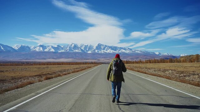 Person walks alone on long straight road stretching toward distant snowy mountains. Clear blue sky with scattered clouds above. Golden-brown grasses line both sides of asphalt path