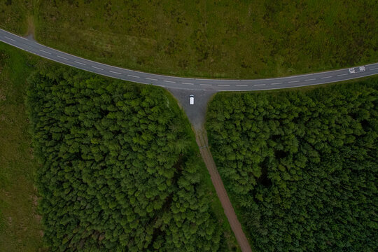 Aerial view of a white vehicle parked in a gravel pull-off area along a winding road cutting through the dense evergreen trees of Penllyn Forest in Bala, Wales, United Kingdom.