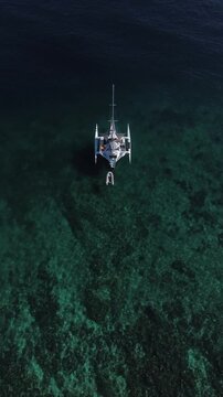 Aerial drone view of a white trimaran sailing yacht anchored on crystal clear turquoise water above a coral reef in the open sea.