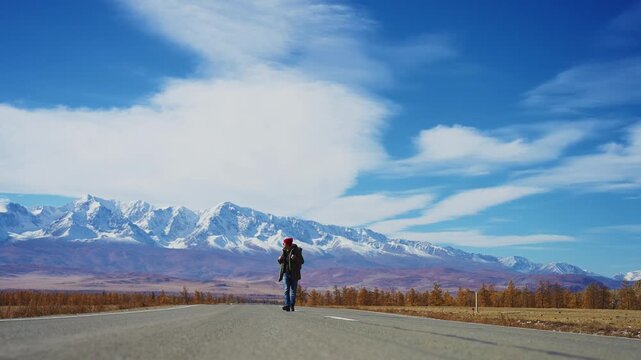 Lone traveler walks down a wide open road toward distant snow-capped mountains. The sky is vast and blue with wispy clouds. Autumn foliage lines the roadside. The person appears calm, focused