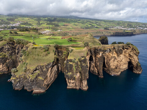 Aerial view of Miradouro do Sertao featuring dramatic volcanic cliffs, lush green fields, and the Atlantic Ocean coastline in Capelas, Acores, Portugal.