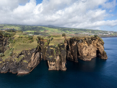 Aerial view of the dramatic coastal cliffs at Miradouro do Sertao, featuring rugged rock formations, lush green fields, and the deep blue Atlantic Ocean in Capelas, Acores, Portugal.