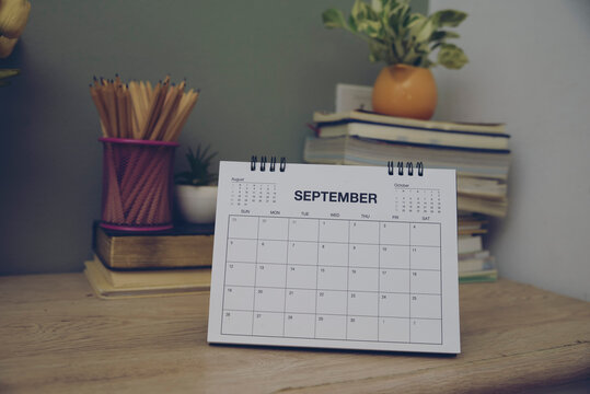 2026 Calendar on wooden desk in indoor workspace with books, pencil holder and flowers for monthly planning, scheduling and organization.