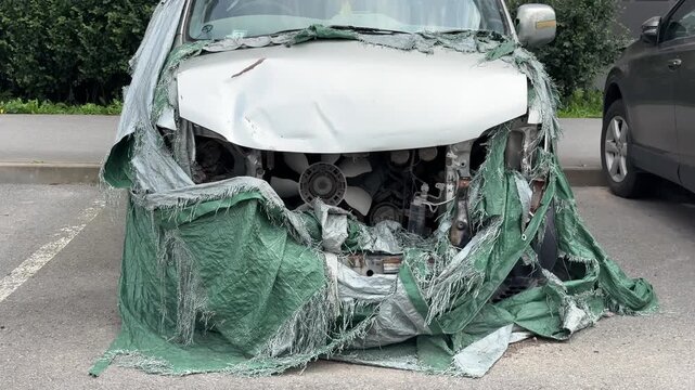 A heavily crashed white car with a severely damaged front structure and a torn green cover is parked in a parking lot, vehicle insurance claim, auto body repair shop, transport safety education.