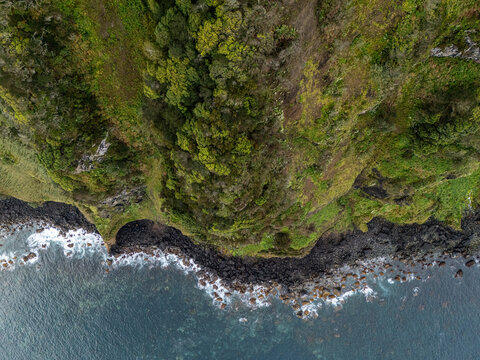 Aerial view of Miradouro de Santa Iria, featuring steep green cliffs, dark volcanic rocks, and crashing waves along the Atlantic Ocean in Porto Formoso, Acores, Portugal.