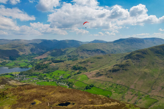 Aerial view of a paraglider soaring over the mountainous landscape of Sheffield Pike with green fields, stone walls, and a lake under a blue sky in Penrith, England, United Kingdom.