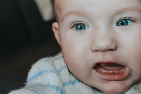 Close-up of a baby with blue eyes wearing a knitted sweater with blue stripes, showing an expressive facial expression against a blurred background