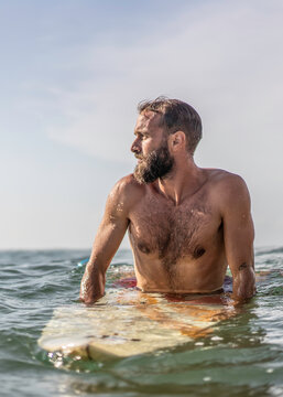Focused millennial surfer sits on his board in the ocean, watching the horizon before the next wave in bright sunlight. Vertical male portrait expresses anticipation, awareness, and resilience.