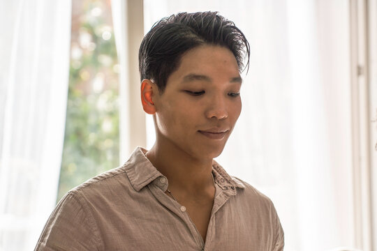 Young asian man in a calm indoor setting with eyes lowered in a quiet mindful moment by the window. Natural lifestyle portrait suggesting meditation, inner peace and wellness.