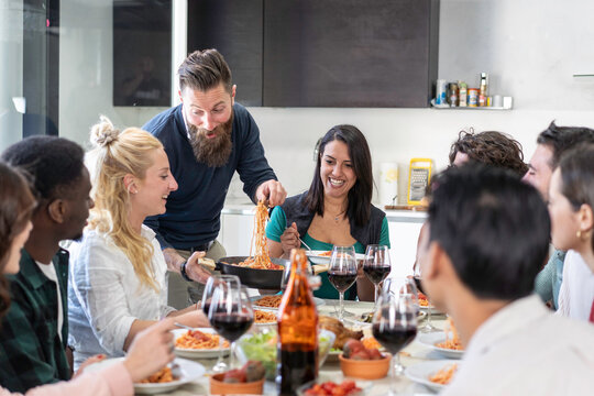 Smiling host serves spaghetti from a pan to diverse friends gathered around a lively dinner table at home. Gen z and millennial adults share homemade food, wine and warm hospitality.
