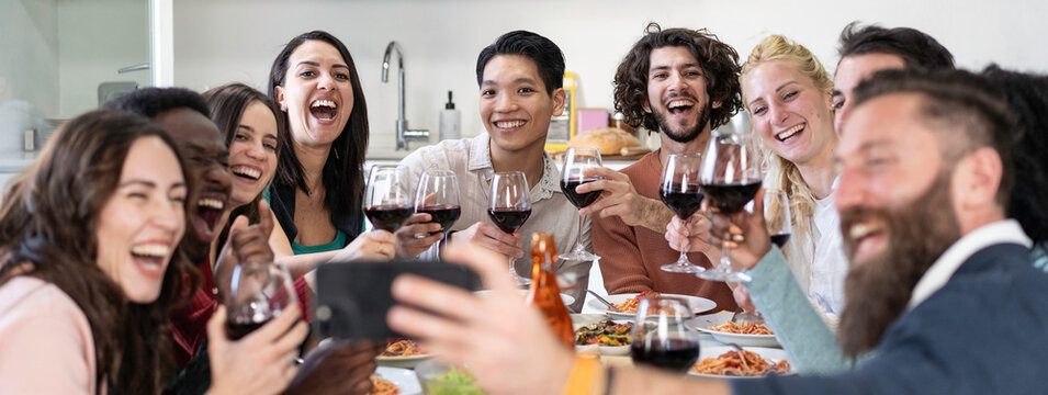Diverse young friends pose for a group selfie while raising red wine glasses during a joyful dinner celebration at home. Gen z and millennial adults capture laughter, friendship and shared memories.