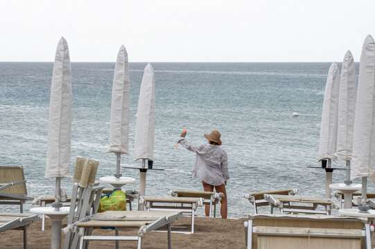 Solo traveler faces the sea on an empty beach with closed umbrellas and loungers. Overcast coastal scene suggests off-season vacation, solitude and quiet escape