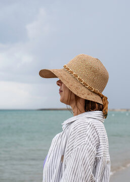 Gen x woman in wide brim sunhat enjoying a peaceful summer day by the sea during a relaxing coastal vacation. Side profile beach portrait with calm water and copy space. Wellness lifestyle concept.