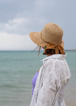 Contemplative woman in straw hat facing the sea beneath a cloudy summer sky. Vertical coastal composition with generous copy space, suited to editorial and book cover use.