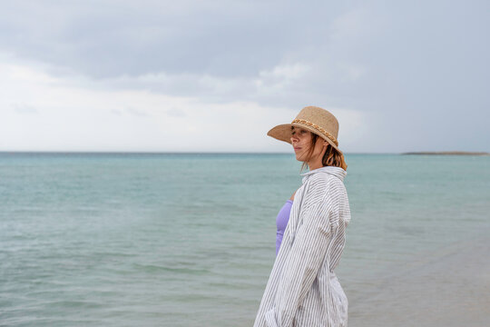 Quiet coastal moment as a gen x woman in a straw hat stands beside turquoise water under a cloudy summer sky. She looks toward the horizon with generous copy space, evoking calm and reflection.