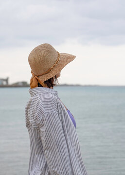 Lost in thought, a gen x woman in a straw hat gazes across the sea beneath a soft overcast sky. Vertical coastal portrait with copy space evokes solitude, travel and reflection.