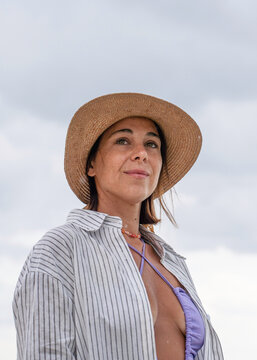 Natural beauty portrait of a confident gen x woman in a straw hat beneath a cloudy summer sky. Vertical composition and soft expression suit wellness, lifestyle and book cover concepts.