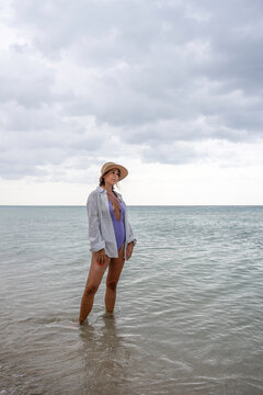 Under a dramatic coastal sky, a gen x woman wades in shallow sea wearing a straw hat and purple swimsuit. Vertical full body scene with copy space suits book covers and travel editorials.