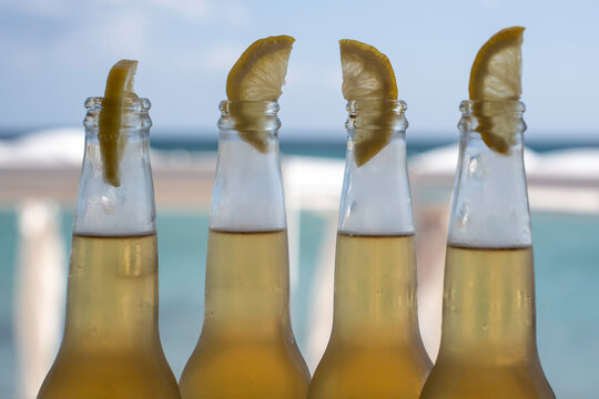Four chilled beer bottles with lemon wedges line up against a soft seaside background on a summer day. Golden drinks and condensation suggest beach bar refreshment and social leisure.