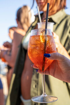 Sunlit spritz cocktail in a woman's hand at a lively beach club. Orange citrus, ice and blue manicure create a stylish summer aperitif scene with friends in soft focus.