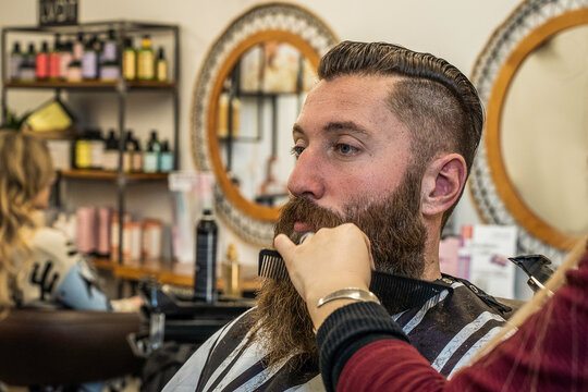 Bearded millennial man receives professional beard combing and shaping in a modern barbershop. Close-up grooming scene highlights precision, masculine style, and personal care.