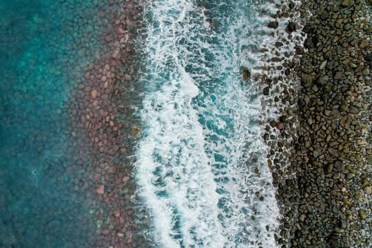 Aerial view of the rocky coastline with turquoise waves crashing against the pebble beach in Ribeira da Janela, Madeira, Portugal.