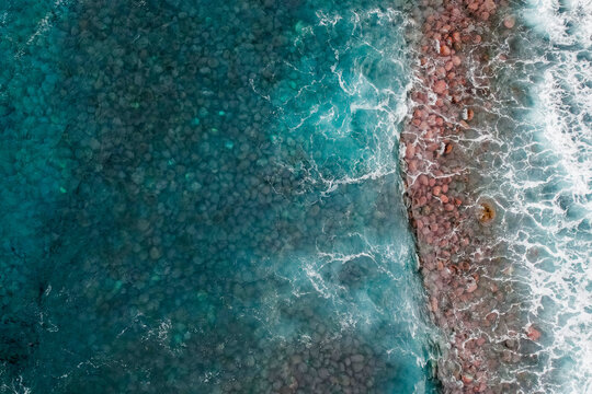 Aerial view of the rocky coastline with turquoise ocean water crashing against reddish-brown pebbles and stones, Ribeira da Janela, Madeira, Portugal.