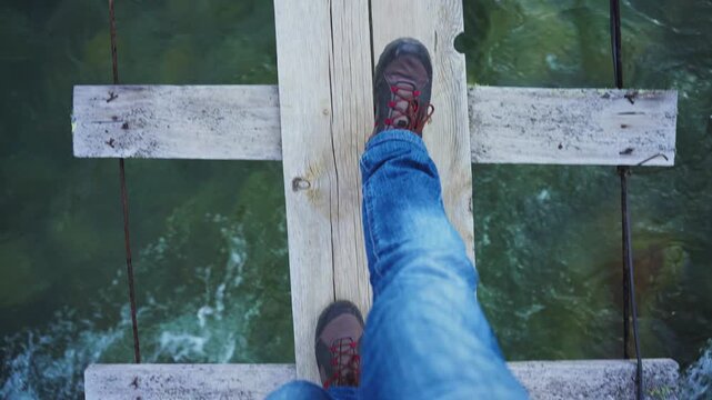 Feet step on weathered wooden bridge over flowing water. View shows worn planks and rushing current below. Person walks carefully across narrow structure. Suggests adventure, nature exploration