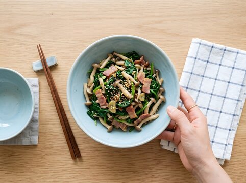 Hand holding ceramic bowl of stir fried spinach with mushrooms and bacon on wooden table, mushroom