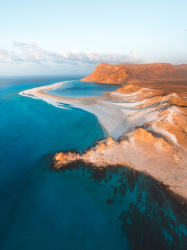 Aerial view of Detwah Lagoon Ramsar Site with its turquoise waters, white sandbar, and rugged coastal cliffs under a clear sky in Socotra, Yemen.