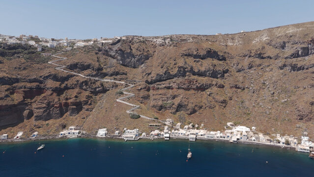 Aerial view of the village of Manolas perched on the volcanic cliffs of Thirasia with a winding zigzag path leading down to the blue sea in Manolas, Greece.