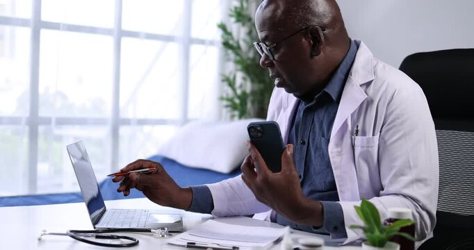 Senior african american male doctor in a white lab coat using a laptop at his desk, managing patient records or conducting research in a clinic setting