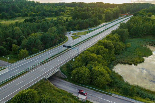Aerial view of a modern highway stretching through a lush green forest landscape with a small pond and a red car driving on a side road in Vilnius, Vilnius County, Lithuania.