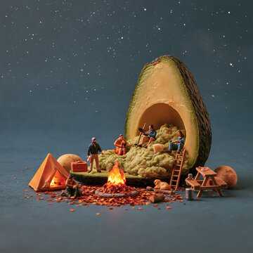 Tiny miniature people are camping and sitting around a small campfire inside a large fresh avocado half on a dark blue background with a starry sky effect