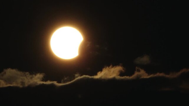 Partial solar eclipse glows in a dark sky as the Moon obscures part of the Sun, while dramatic clouds form a silhouetted layer below.