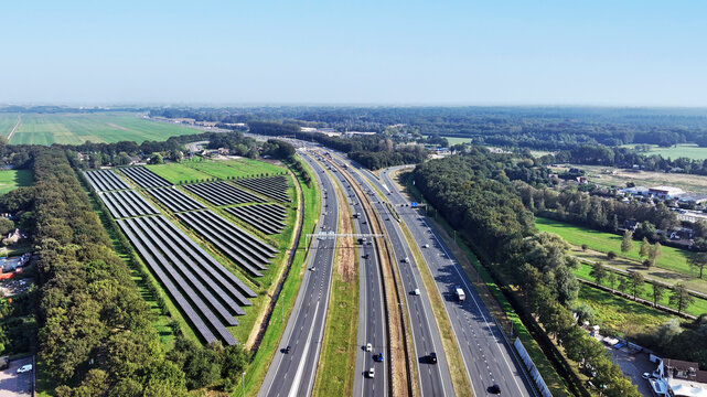 Aerial view of the A1 highway running through the landscape with an adjacent solar farm, green fields, and surrounding forests in Baarn, Utrecht, Netherlands.