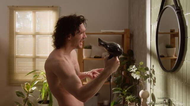 Caucasian young man drying curly hair with hair dryer in bathroom, singing and having fun in front of mirror. Perfect for male grooming, self-care, and lifestyle advertising campaigns