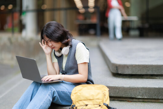 Young asian female student sits on outdoor stairs, appearing frustrated and tired while working on her laptop, rubbing her eye, indicating stress and mental fatigue from studying