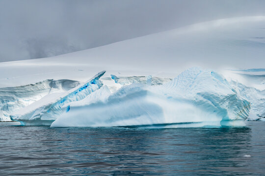Aerial view of a large iceberg floating in the cold waters with a vast snow-covered glacier and cloudy sky in the background in Antarctica.