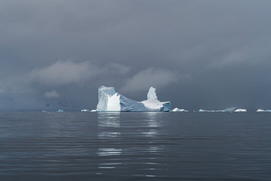 Aerial view of a large iceberg floating in the calm dark waters of the Southern Ocean under a dramatic cloudy sky in Antarctica.