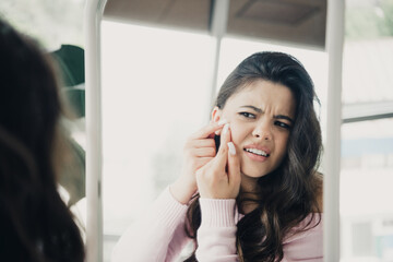 Obraz premium Young woman examining and popping a pimple in the mirror with frustrated expression while performing skincare routine