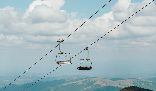 Two empty chairlift seats hang above a mountain landscape in Kopaonik, Serbia. Cable lines cut across the sky over distant hills and forests